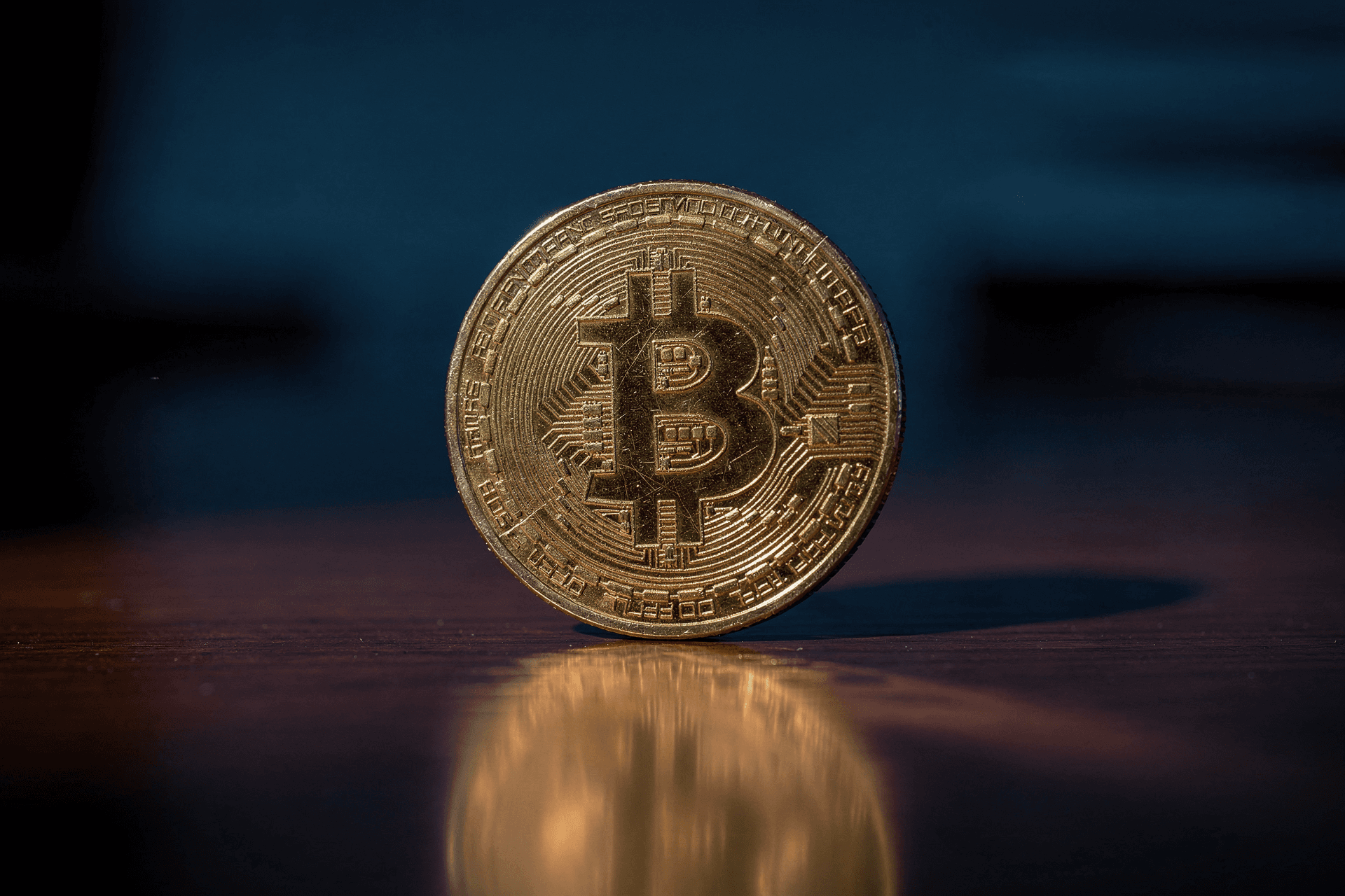 A close-up, illuminated shot of a gold Bitcoin coin standing upright on a reflective wooden surface with a dark background.