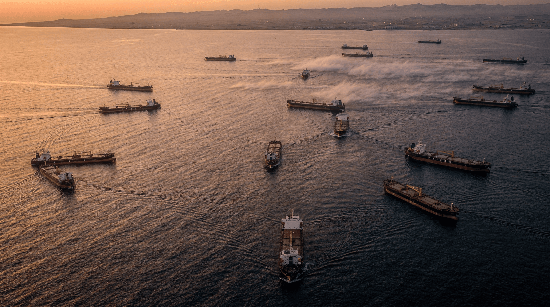 An aerial view of numerous oil tankers sailing on the open sea under a golden-orange sky at dusk, with distant land.