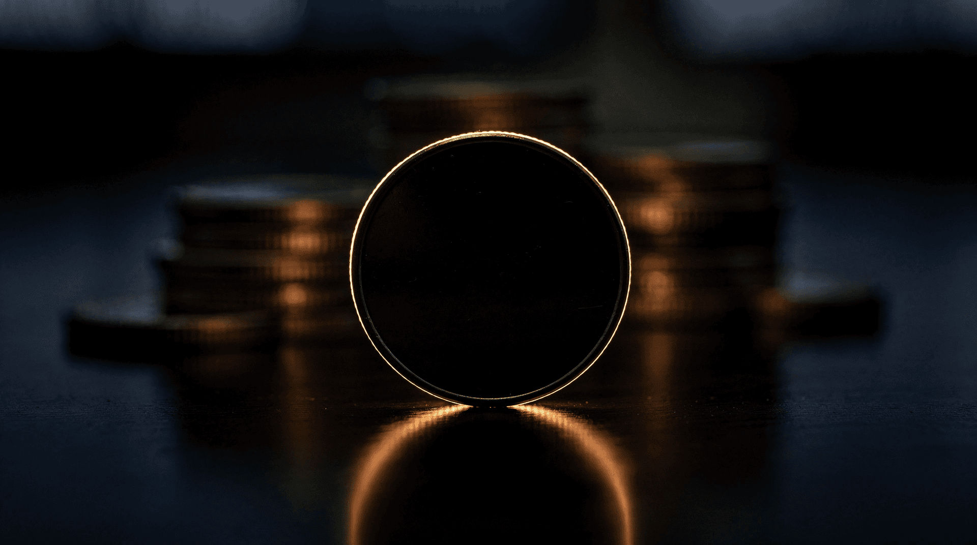 A close-up shot of a dark, generic coin standing upright, reflecting a golden glow onto a dark surface, with blurred coin stacks behind.