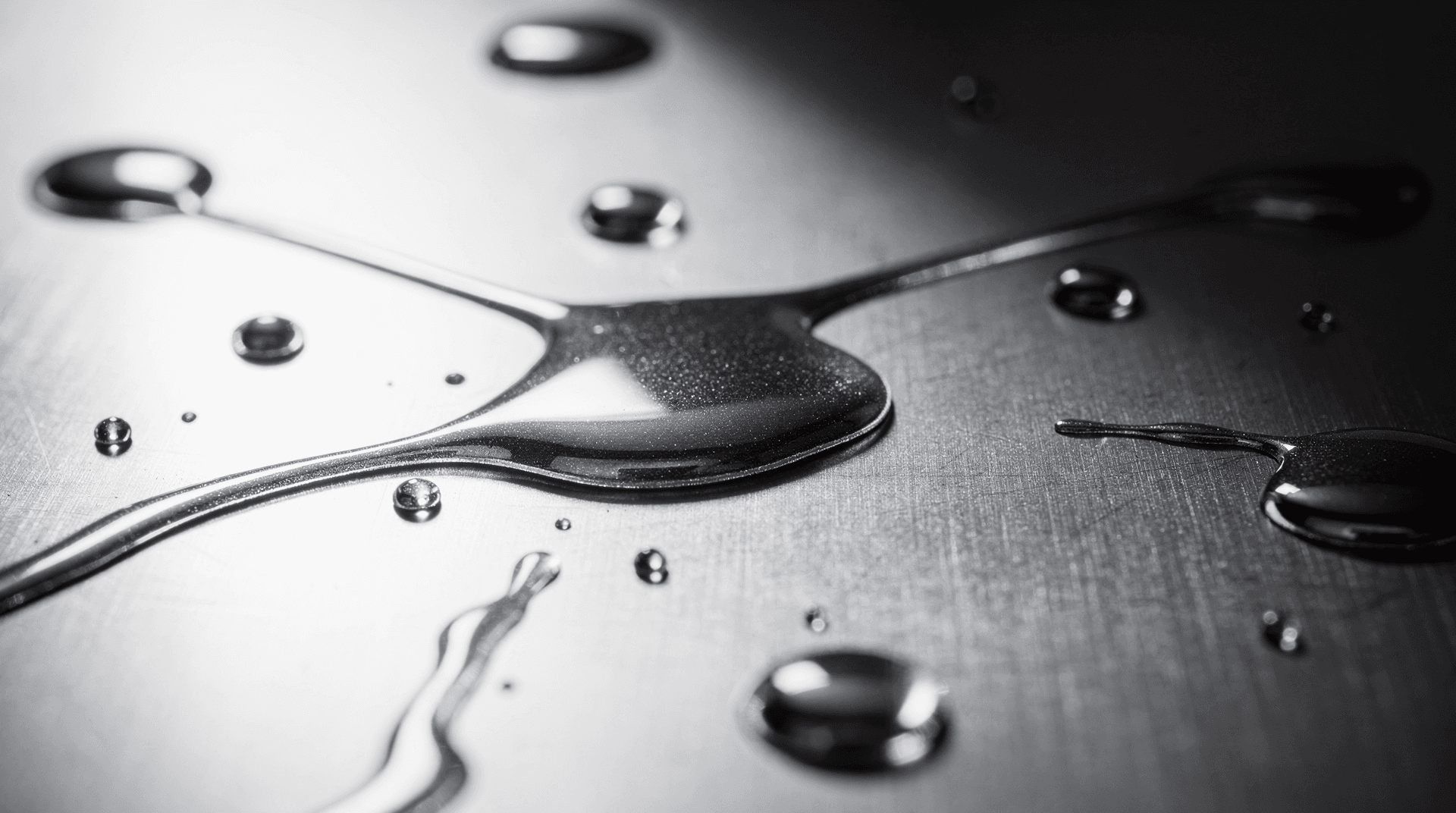 liquid mercury or a similar metallic liquid spilled and forming drops and puddles on a brushed metal surface