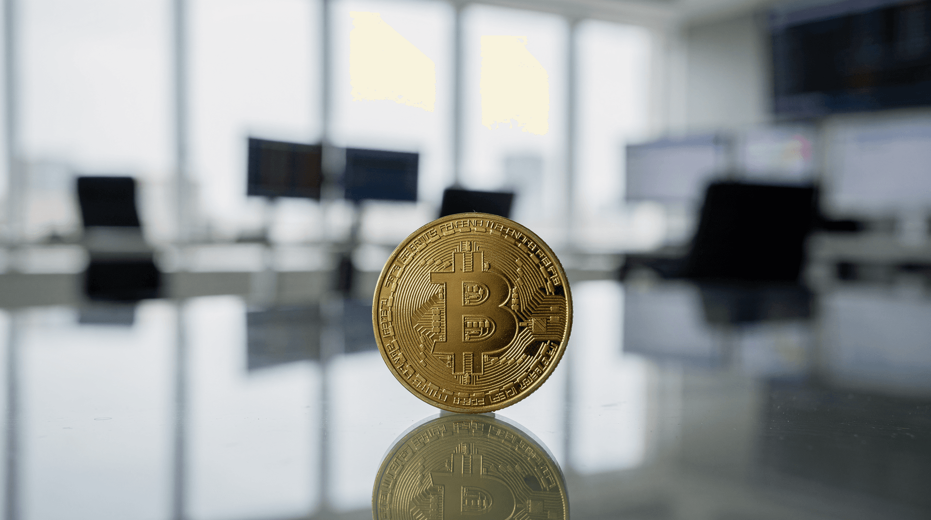 A golden Bitcoin physical coin stands upright on a reflective surface in a modern office with blurred computers in background.