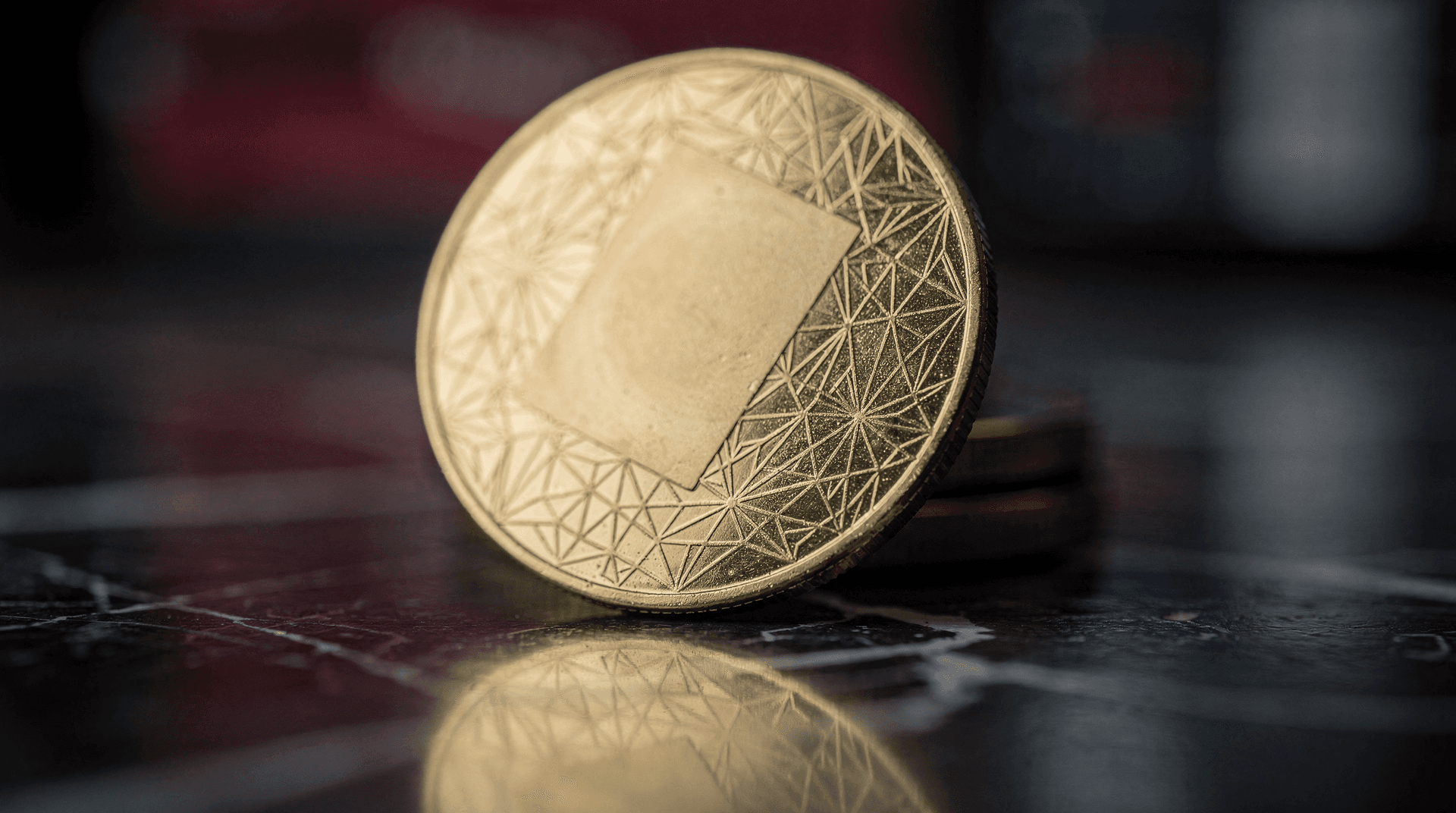 Close-up of a gold-colored coin with a blank square in the center and a geometric pattern around the edge, on a dark reflective surface