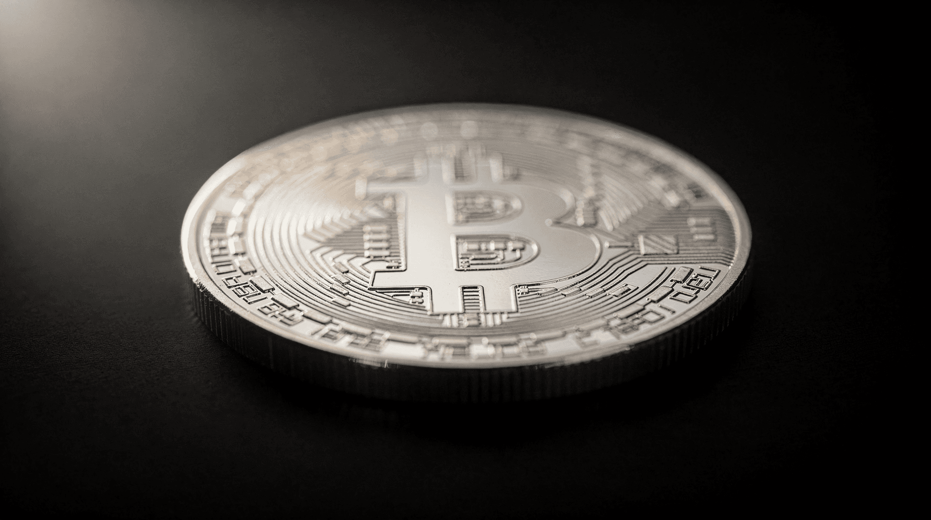 A silver-colored physical Bitcoin coin resting on a dark surface, with soft light illuminating its top left portion.