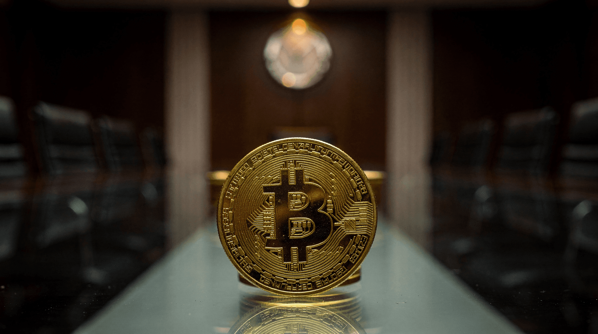 A golden Bitcoin coin standing upright on a glass conference table in a dimly lit boardroom, chairs blurred in background.