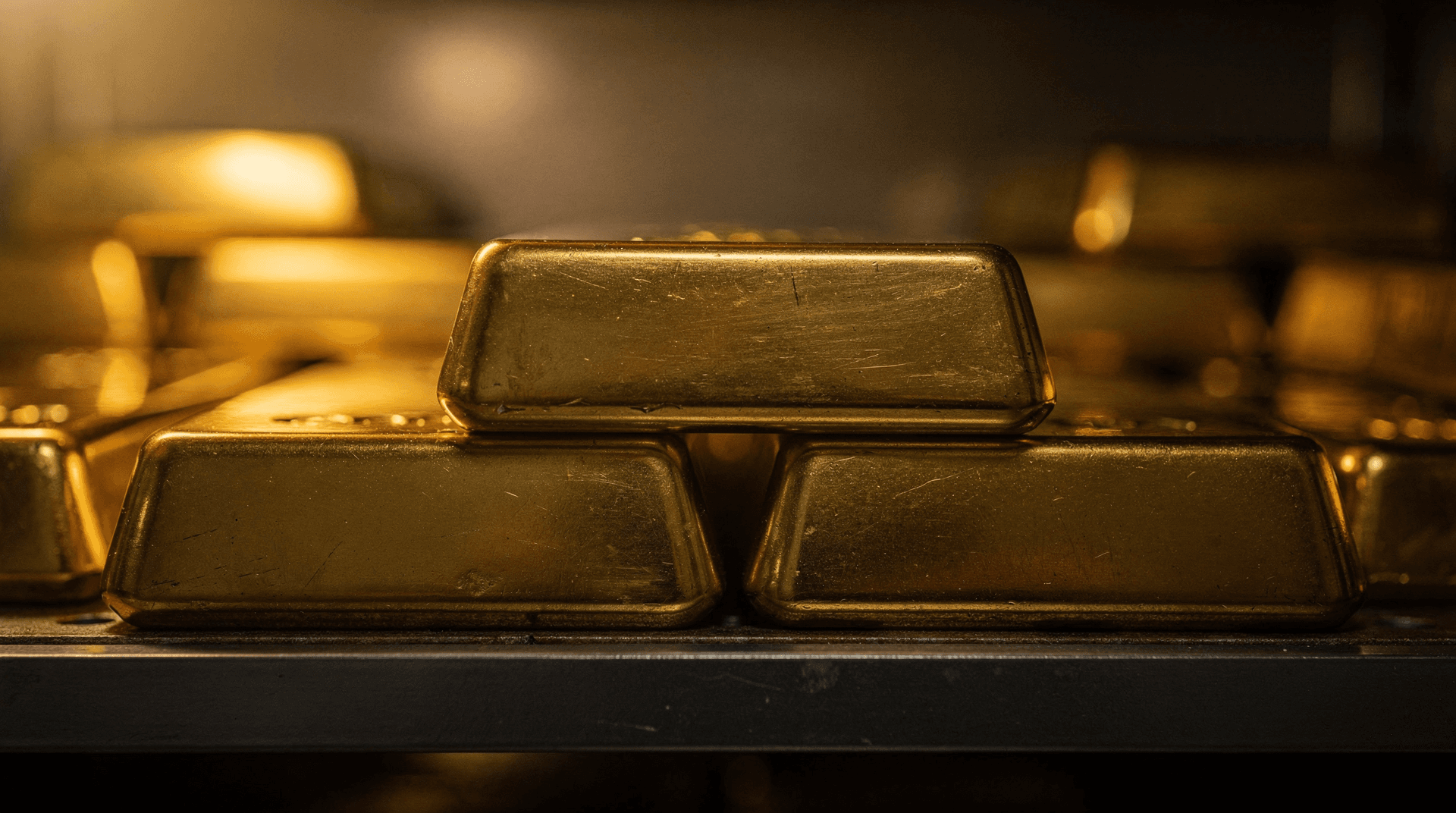 Close-up of several shiny, stacked gold bars on a dark shelf, with more gold in the blurred background.