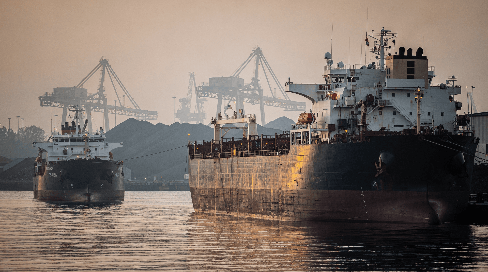 A large bulk carrier looms over a smaller one anchored nearby in a hazy industrial harbor at dusk.