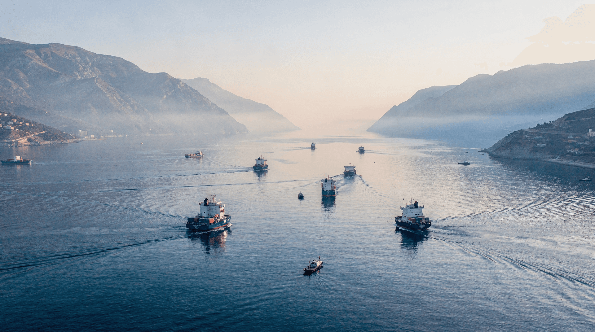 Aerial view of a narrow strait with numerous cargo ships and smaller boats navigating the calm waters between misty mountains.