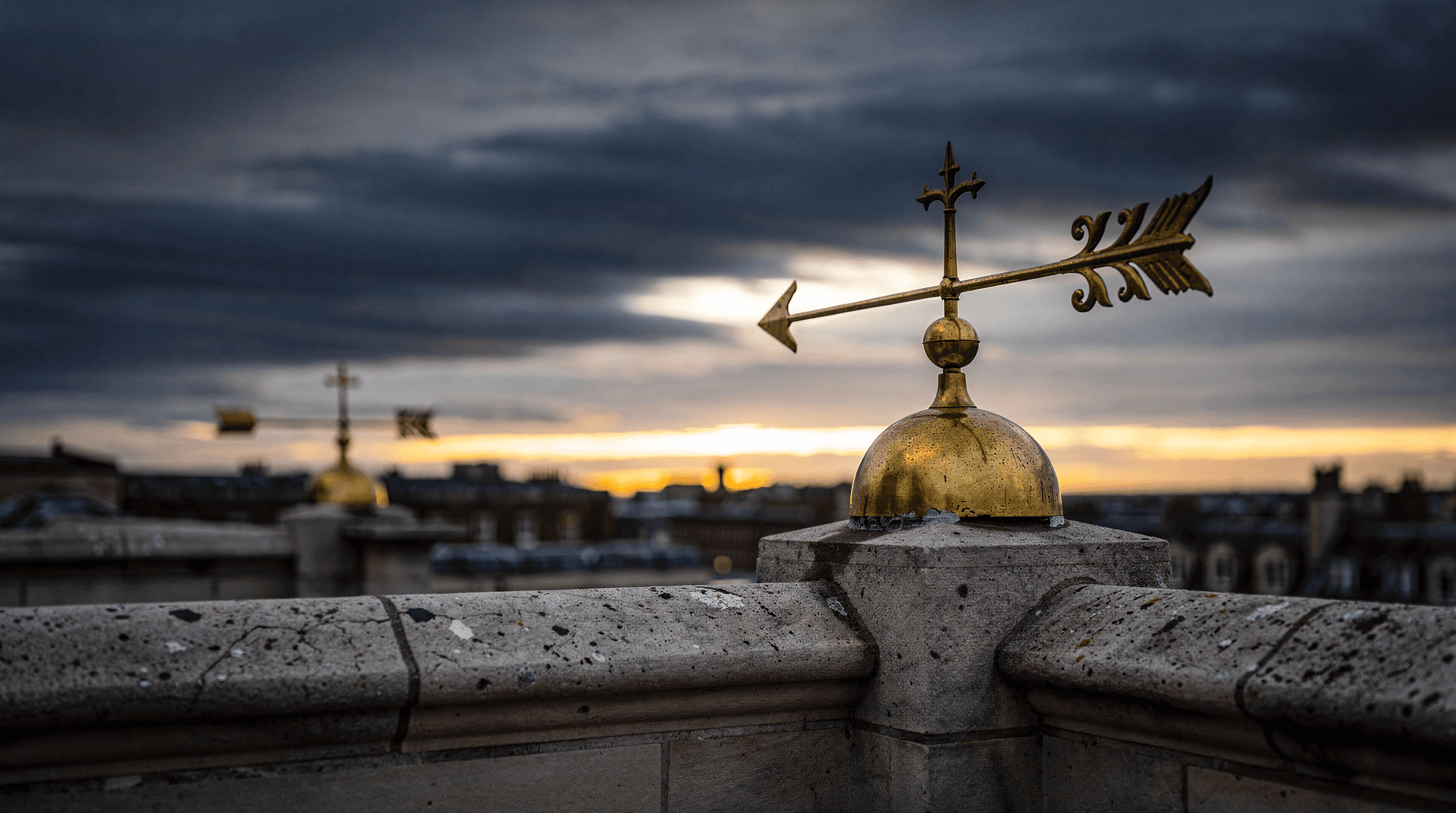 A brass weather vane atop a stone rooftop swings hard against a fast-moving stormy dusk sky.