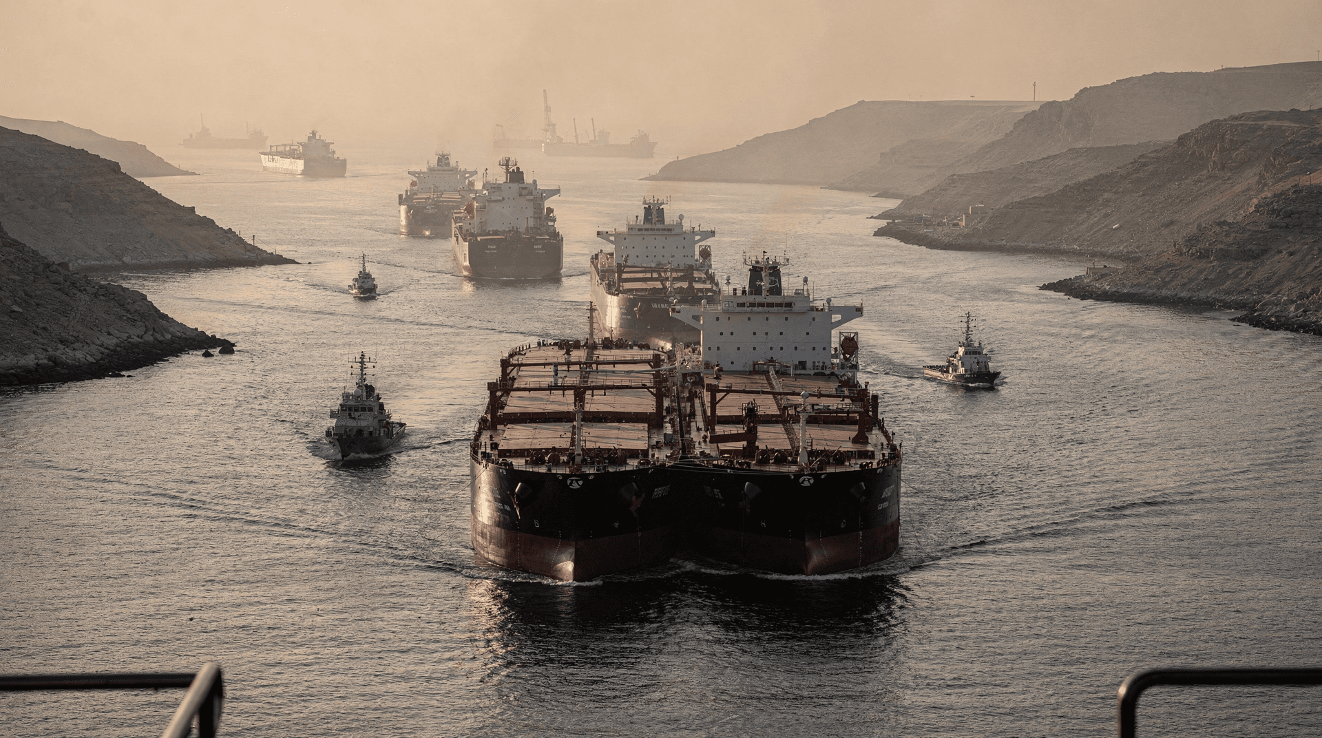 A convoy of massive oil tankers and smaller pilot boats navigate a narrow strait with arid, rocky hillsides under a hazy sky.