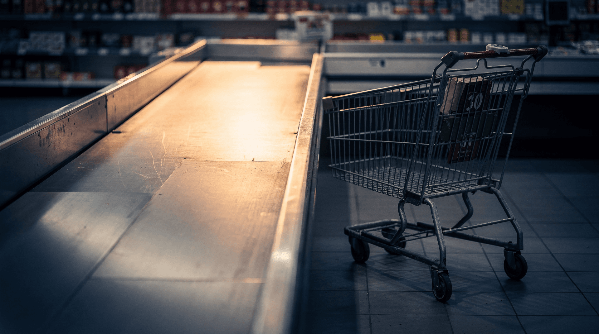 An empty grocery checkout lane with a bare conveyor belt lit in warm gold light against deep shadow