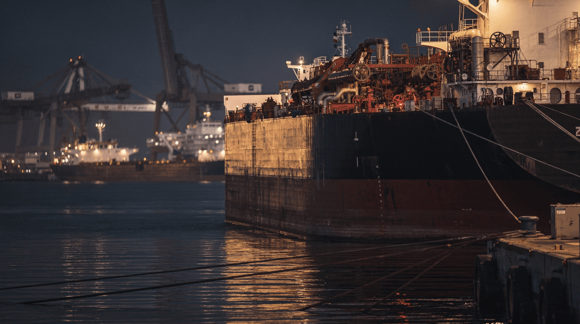 A lone oil tanker eases away from a Gulf port dock at dusk while other vessels remain moored under cranes.