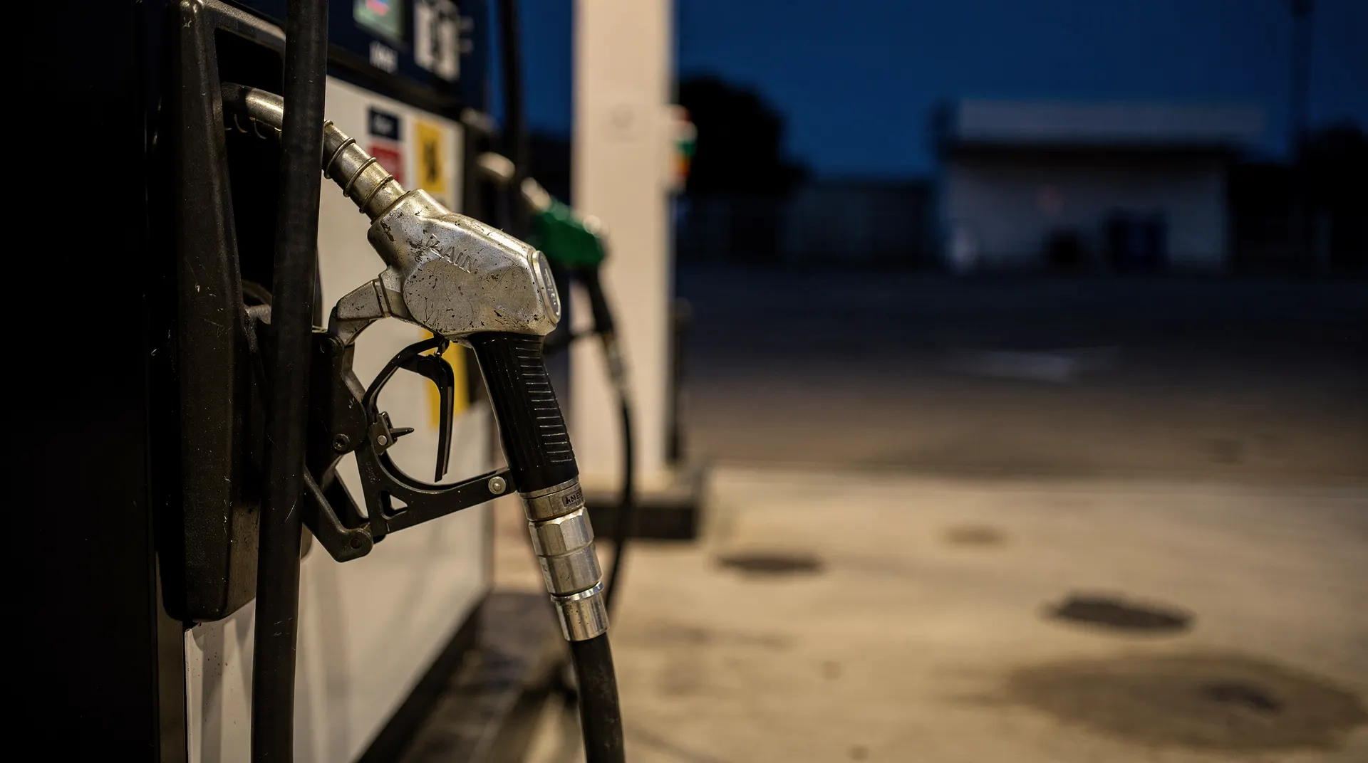 A fuel-pump nozzle hangs at an empty forecourt at dusk, with the pump housing blurred behind it.