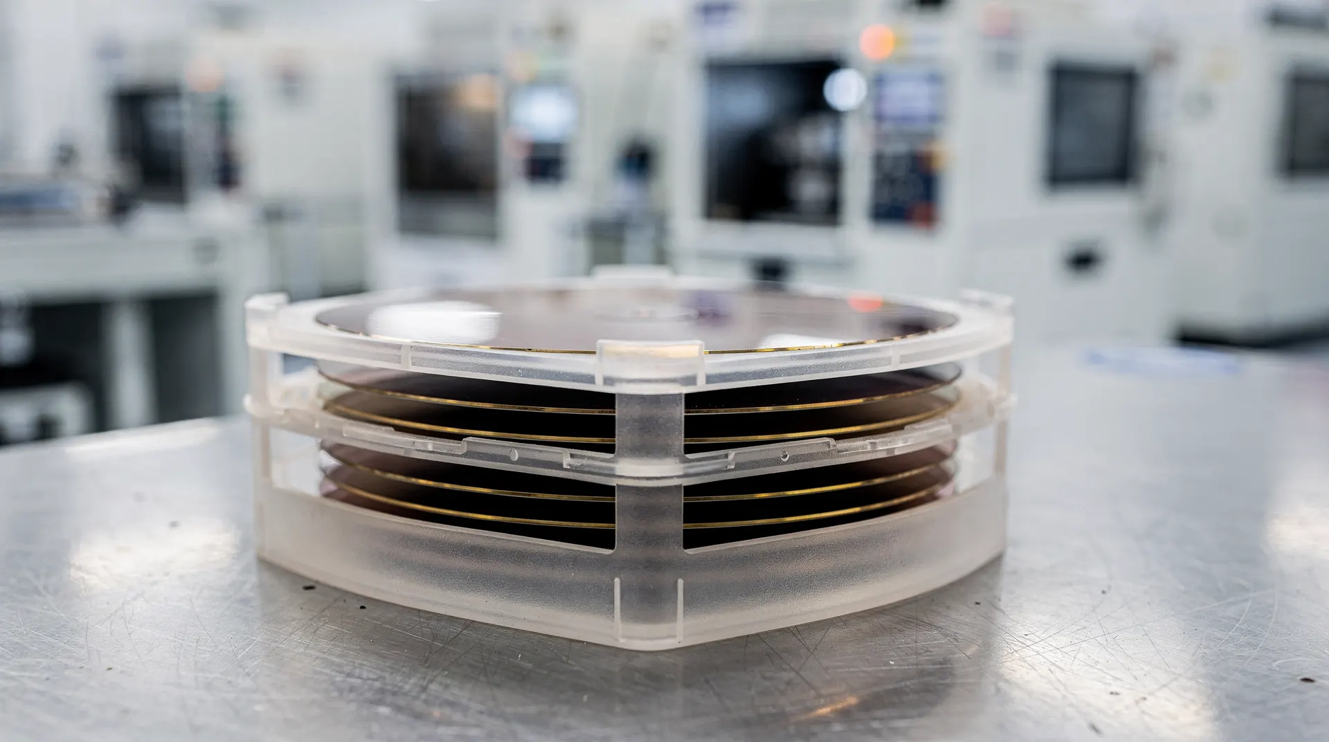 Silicon wafers stand in a carrier, with semiconductor fabrication equipment blurred behind them.