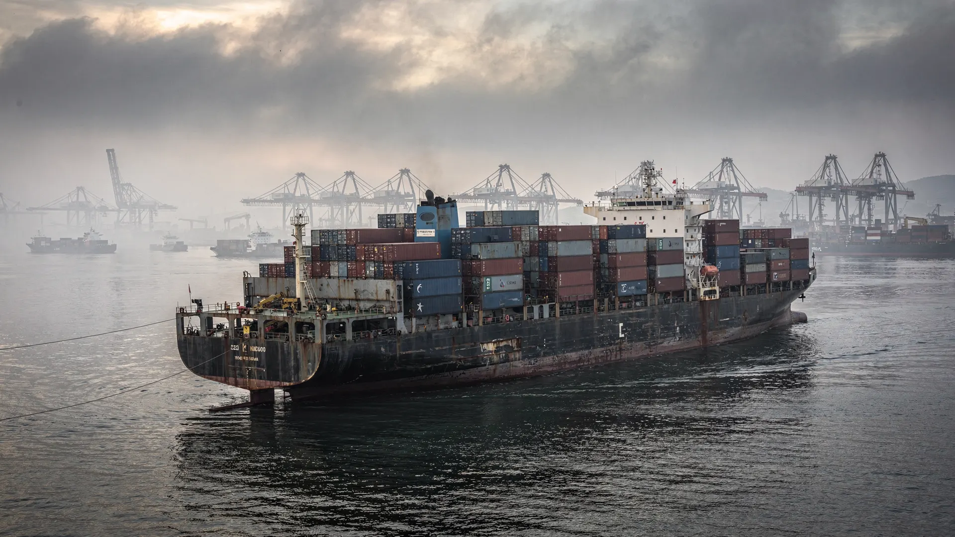 Container ships wait at anchor in a hazy harbor, with cargo cranes in the distance.