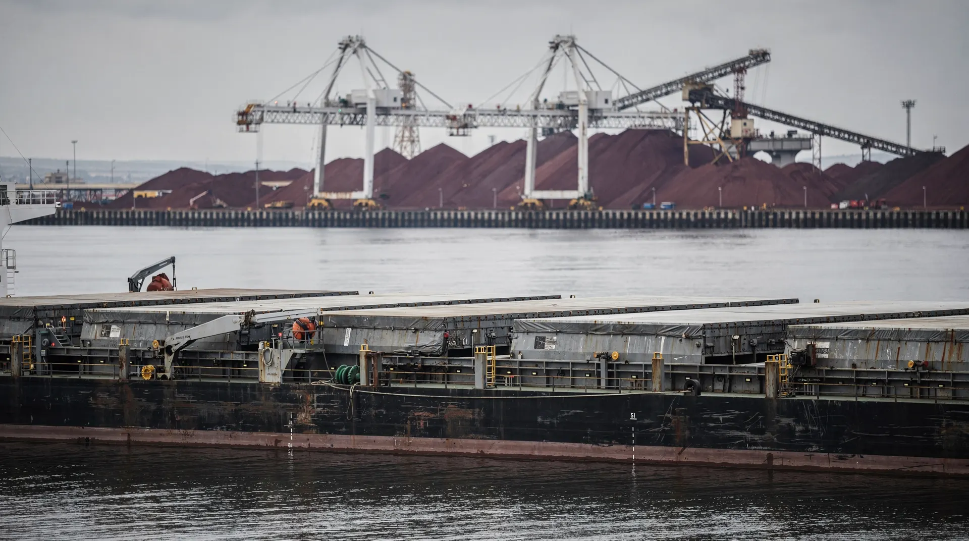 A Capesize dry-bulk carrier rests on calm water near distant iron-ore loading cranes under an overcast sky.