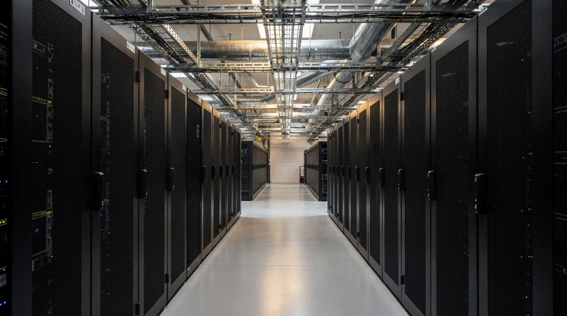 Closed server racks recede down a dim data-center aisle beneath overhead cooling pipes.