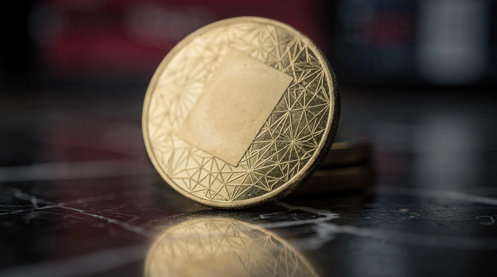Close-up of a gold-colored coin with a blank square in the center and a geometric pattern around the edge, on a dark reflective surface