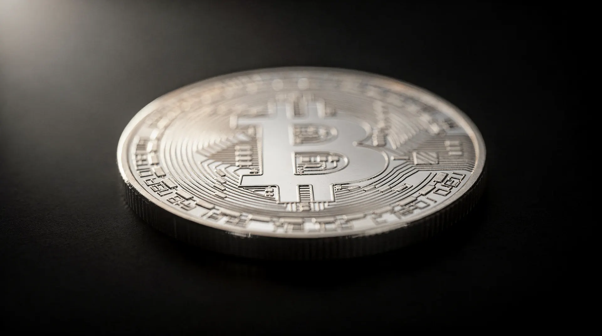 A silver-colored physical Bitcoin coin resting on a dark surface, with soft light illuminating its top left portion.