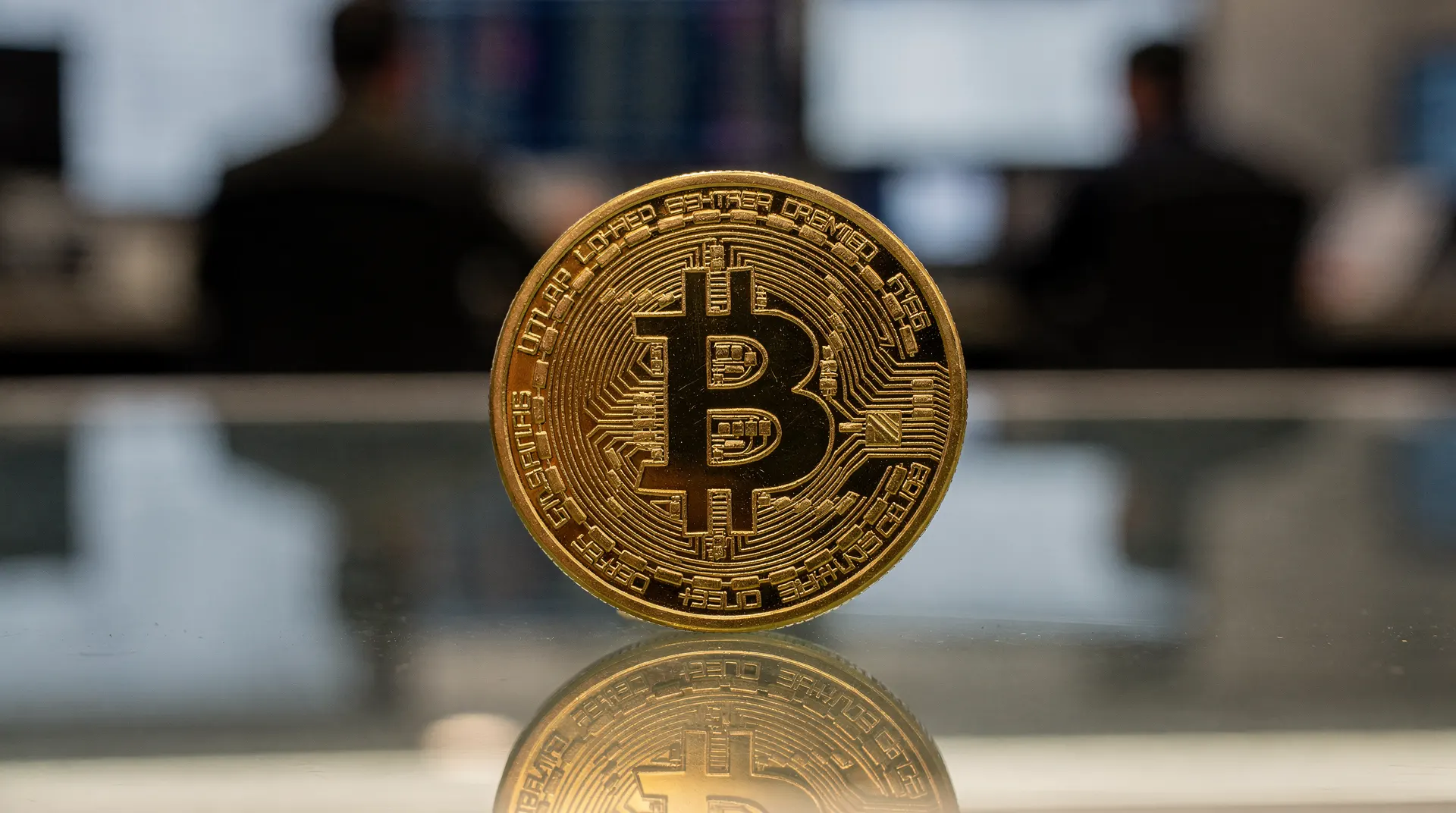 A golden physical Bitcoin coin stands upright on a reflective surface, with two blurred figures in suits in the background.
