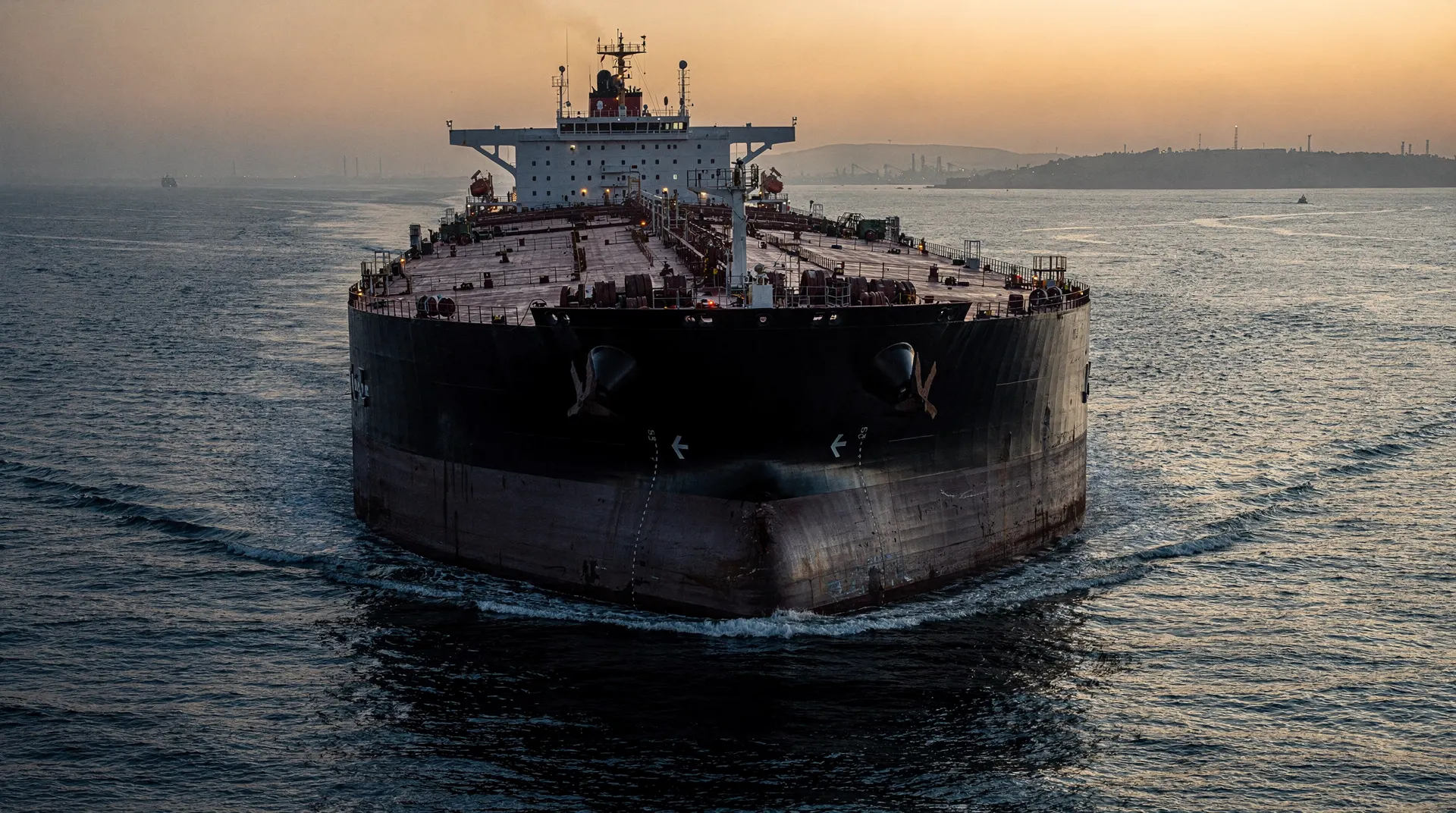 An unmarked crude oil tanker crosses a narrow sea passage at dusk.