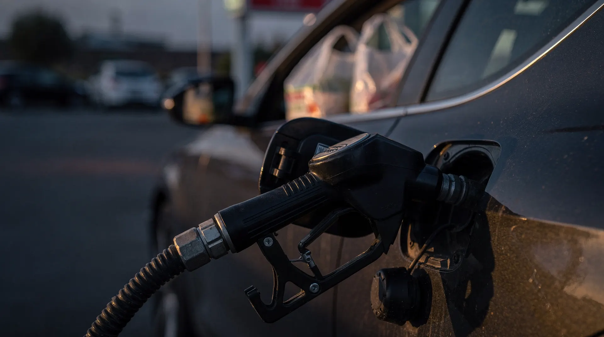 A fuel-pump nozzle rests in a car's fuel port, with grocery bags blurred through the window.