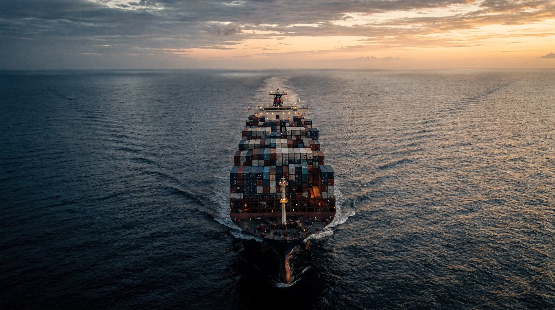 A loaded container ship crosses open water at dusk, leaving a faint wake behind it.