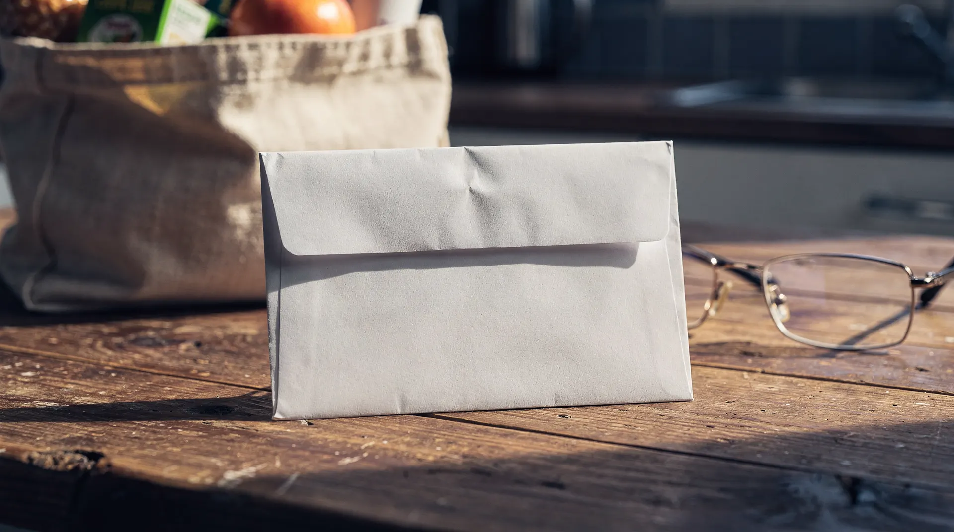 A sealed unmarked envelope rests on a kitchen table beside reading glasses and a grocery bag.