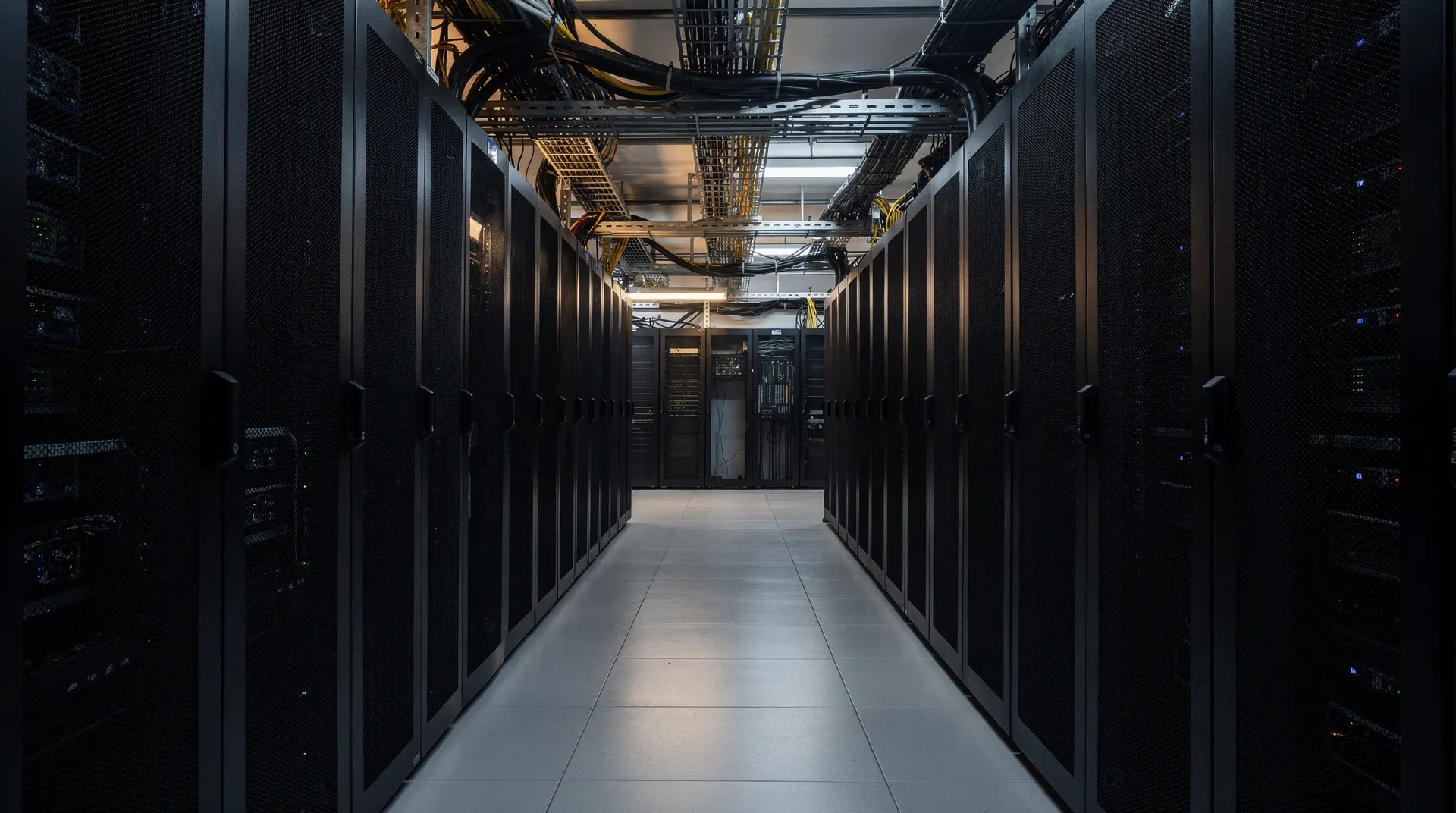 Empty data-center aisle lined with server racks and heavy power conduits, fading into shadow.