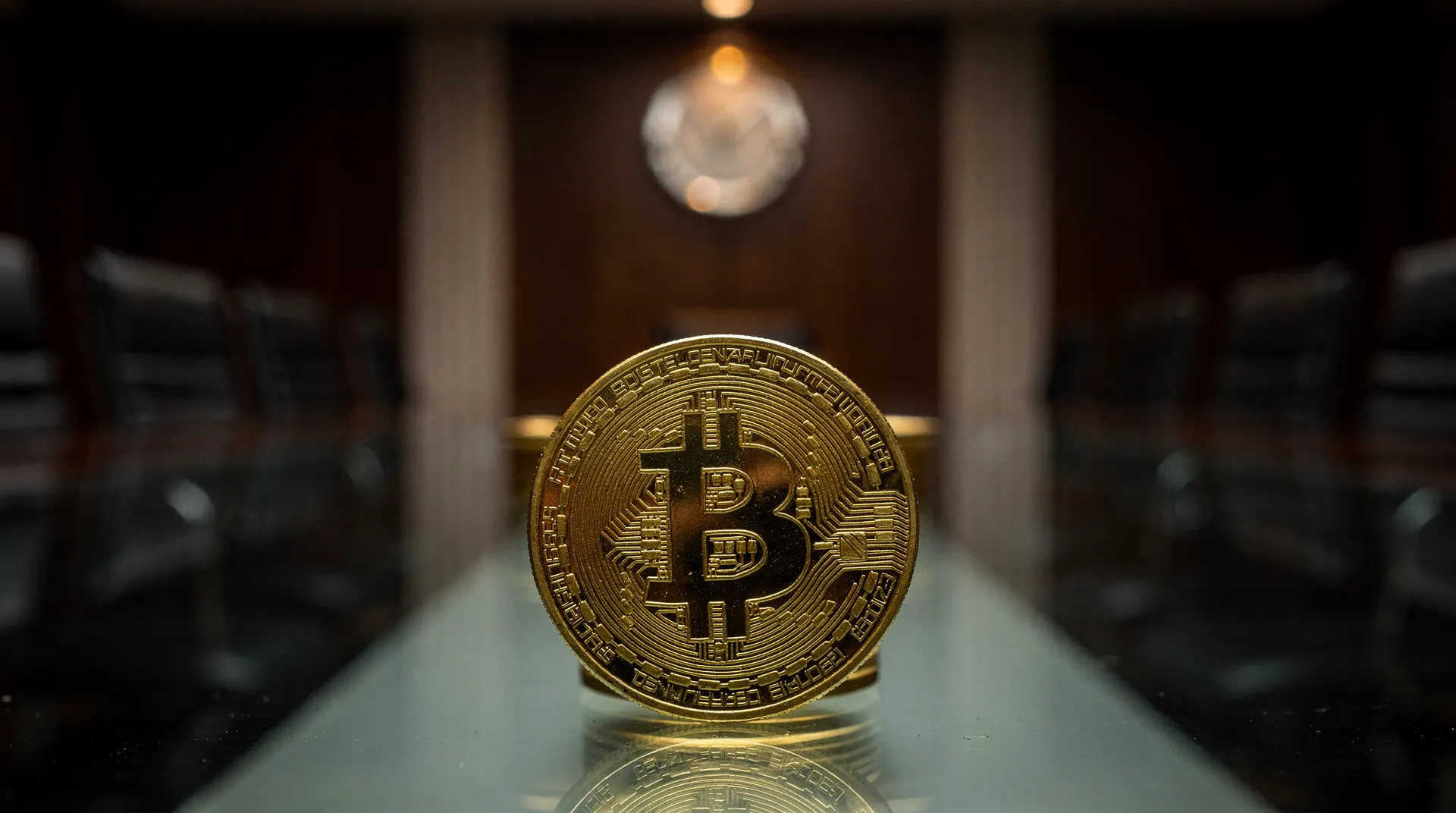 A golden Bitcoin coin standing upright on a glass conference table in a dimly lit boardroom, chairs blurred in background.