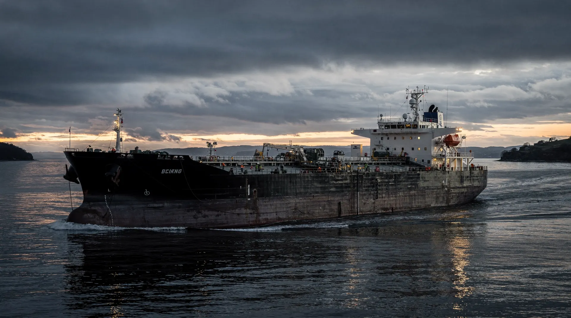 An oil tanker passes between distant coastlines beneath dark clouds at dusk.