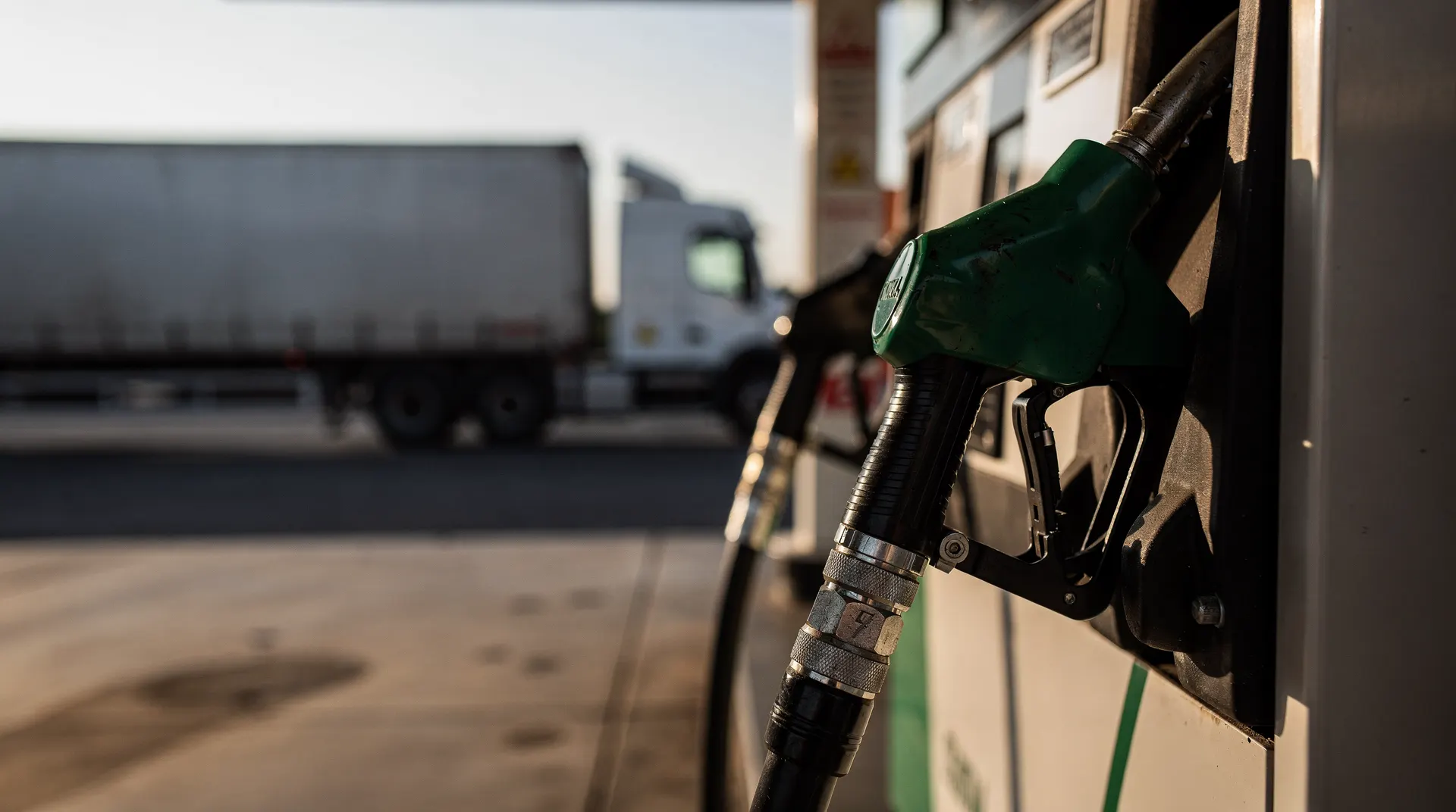 A diesel nozzle hangs in the foreground of a quiet forecourt, with another nozzle and a freight truck blurred behind it.