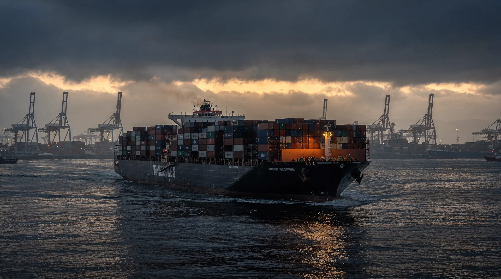 A loaded container ship leaves a hazy port beneath dark clouds at dusk.