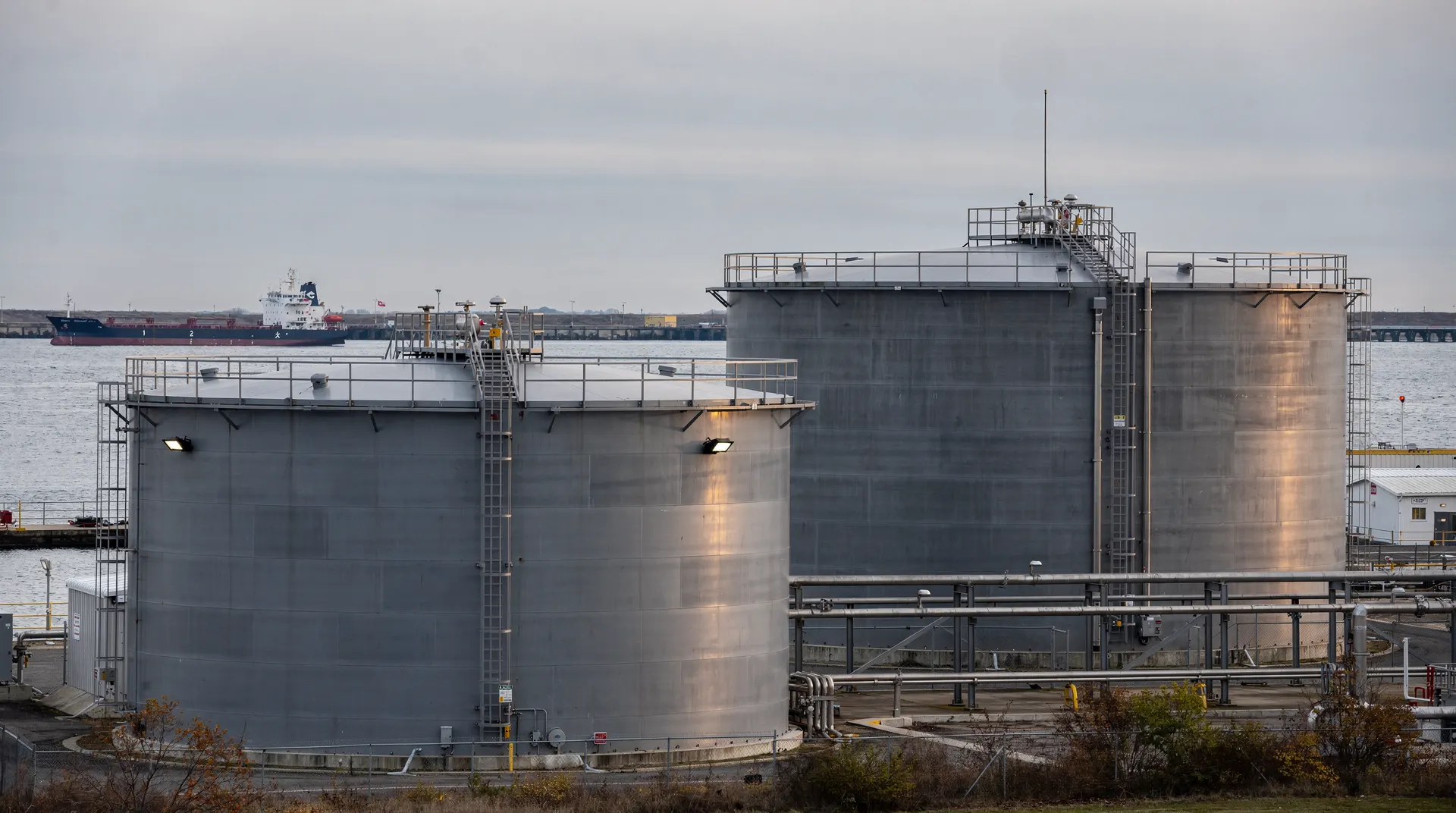Storage tanks at a coastal fuel terminal, with a tanker at a distant berth under an overcast sky.