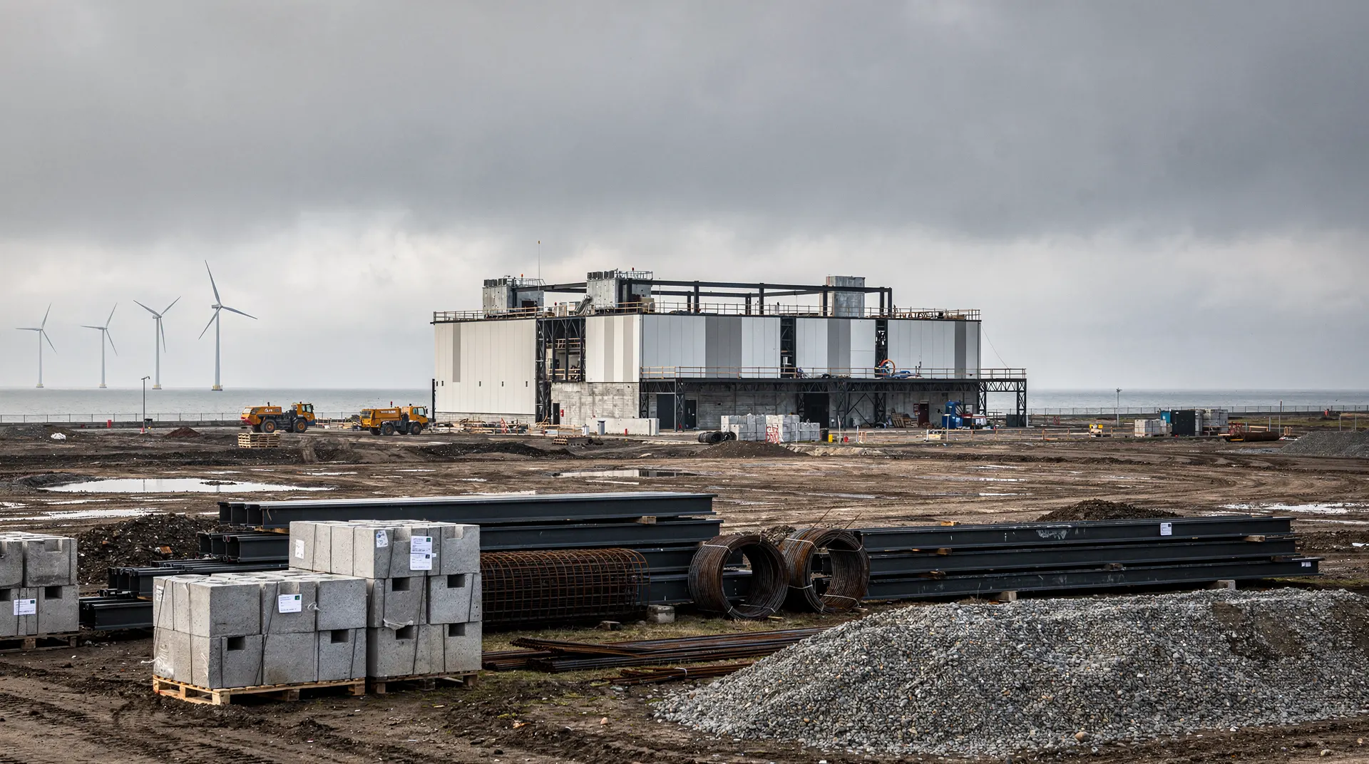 An unfinished data centre building stands beneath an overcast sky, with wind turbines on the distant horizon.