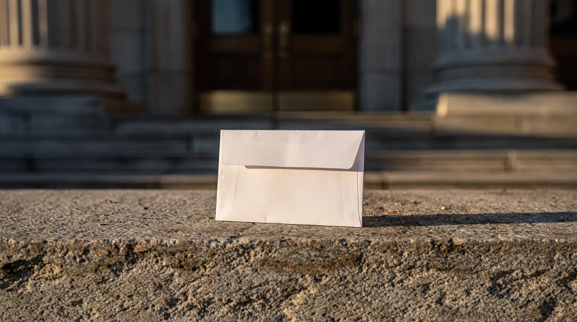 A blank sealed envelope rests on a stone ledge outside an unmarked building entrance.