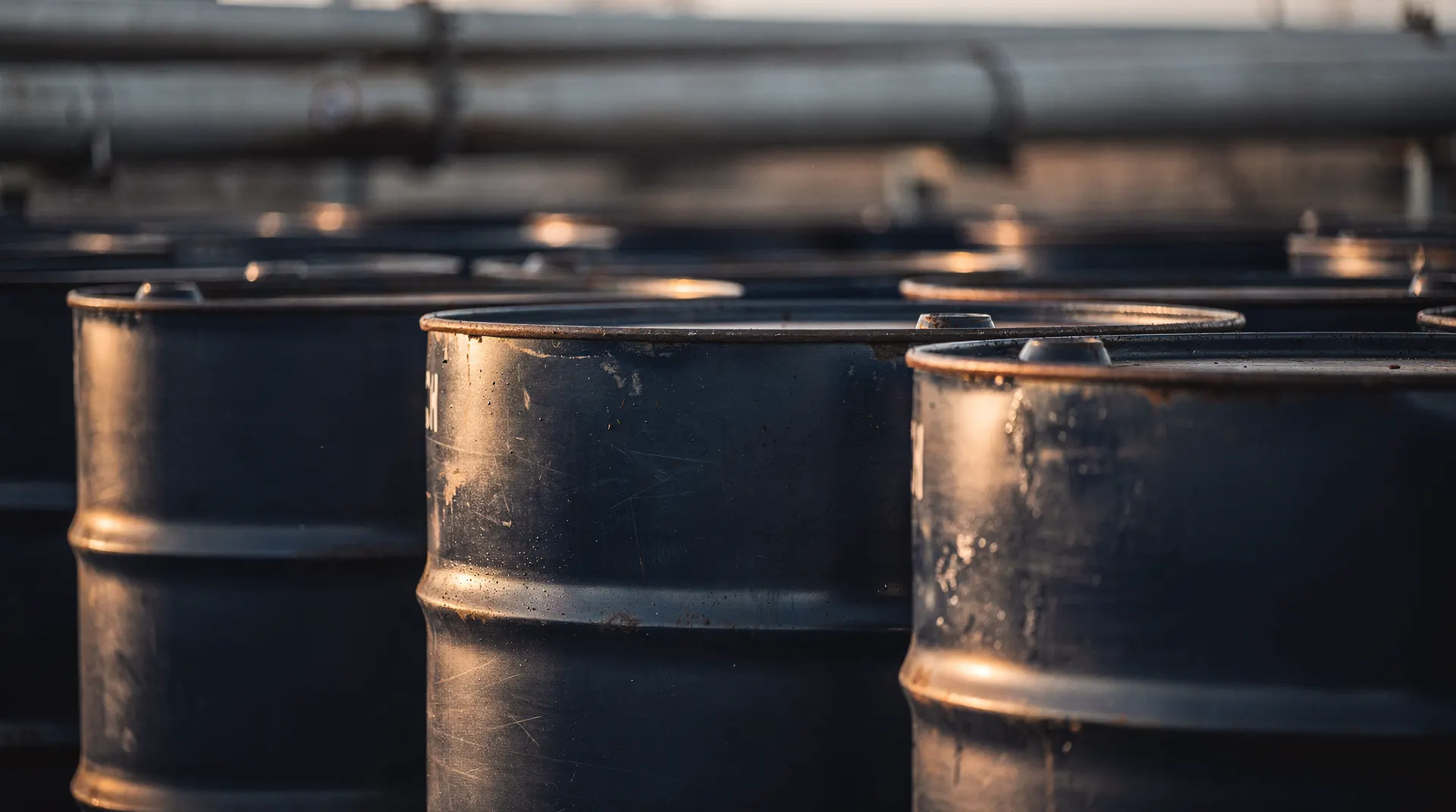 Unmarked oil barrels stand in warm light at an industrial storage site.