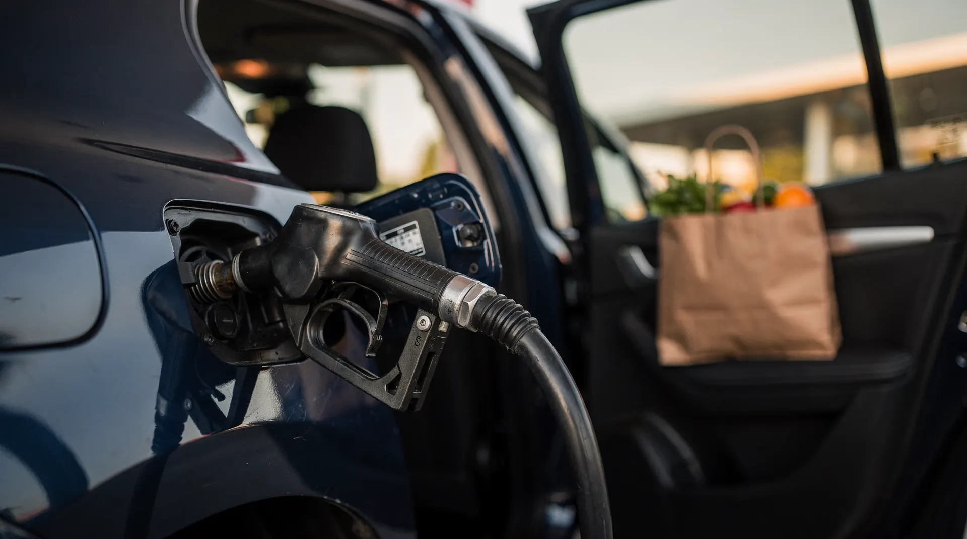 A fuel nozzle fills a car while grocery bags sit inside its open rear door.