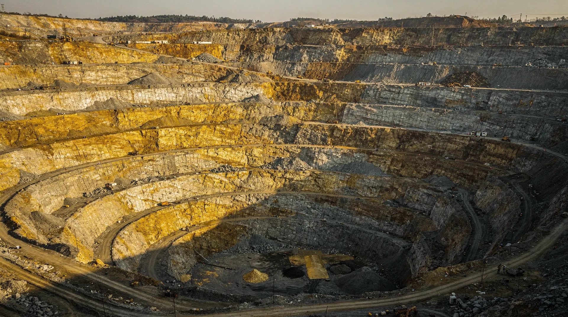 An aerial view of a massive open-pit gold mine, showing terraced layers of rock and earth, with some areas gleaming golden.