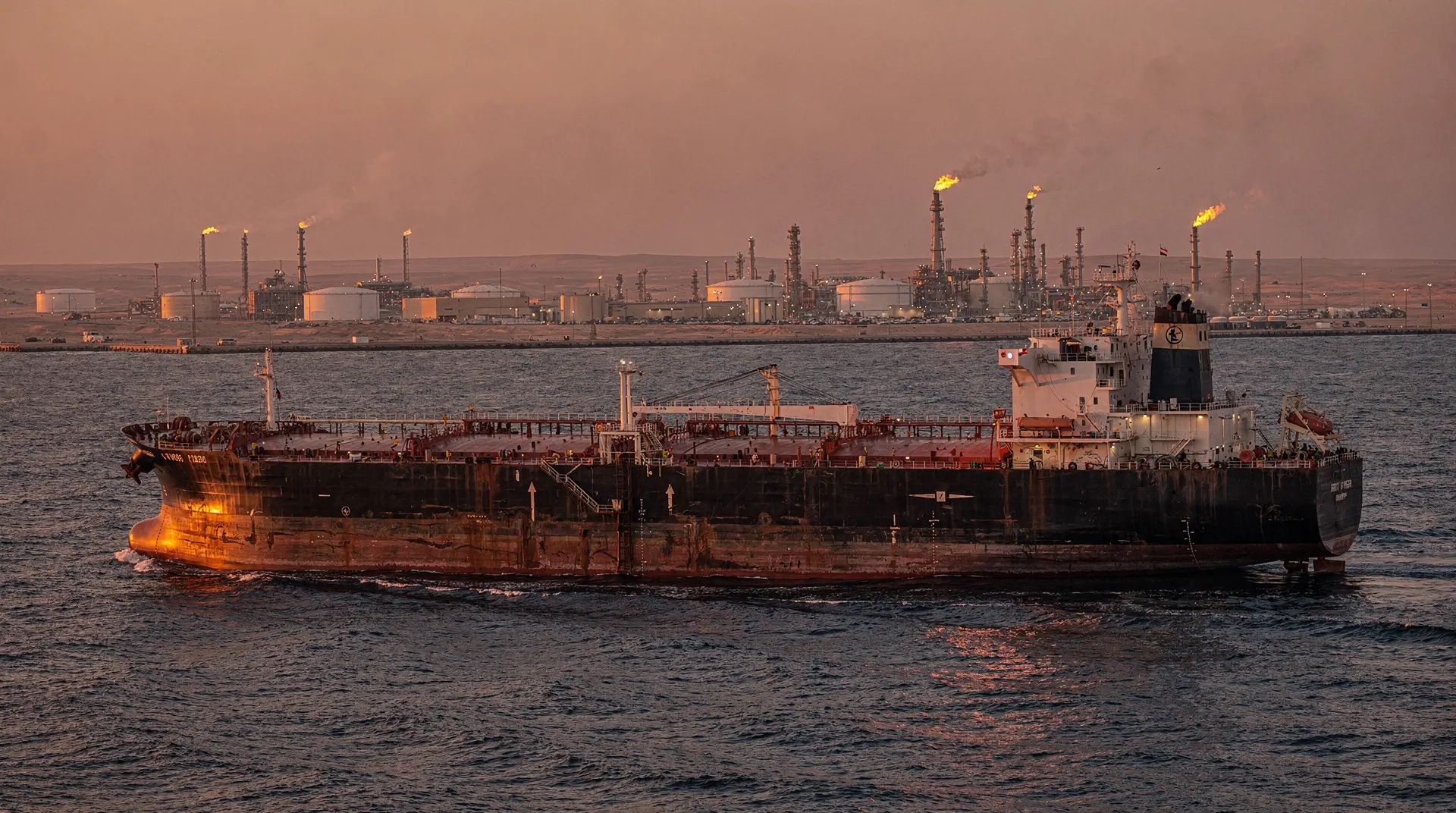 Oil tanker on water with a refining facility and storage tanks in the background under a sunset-colored sky.