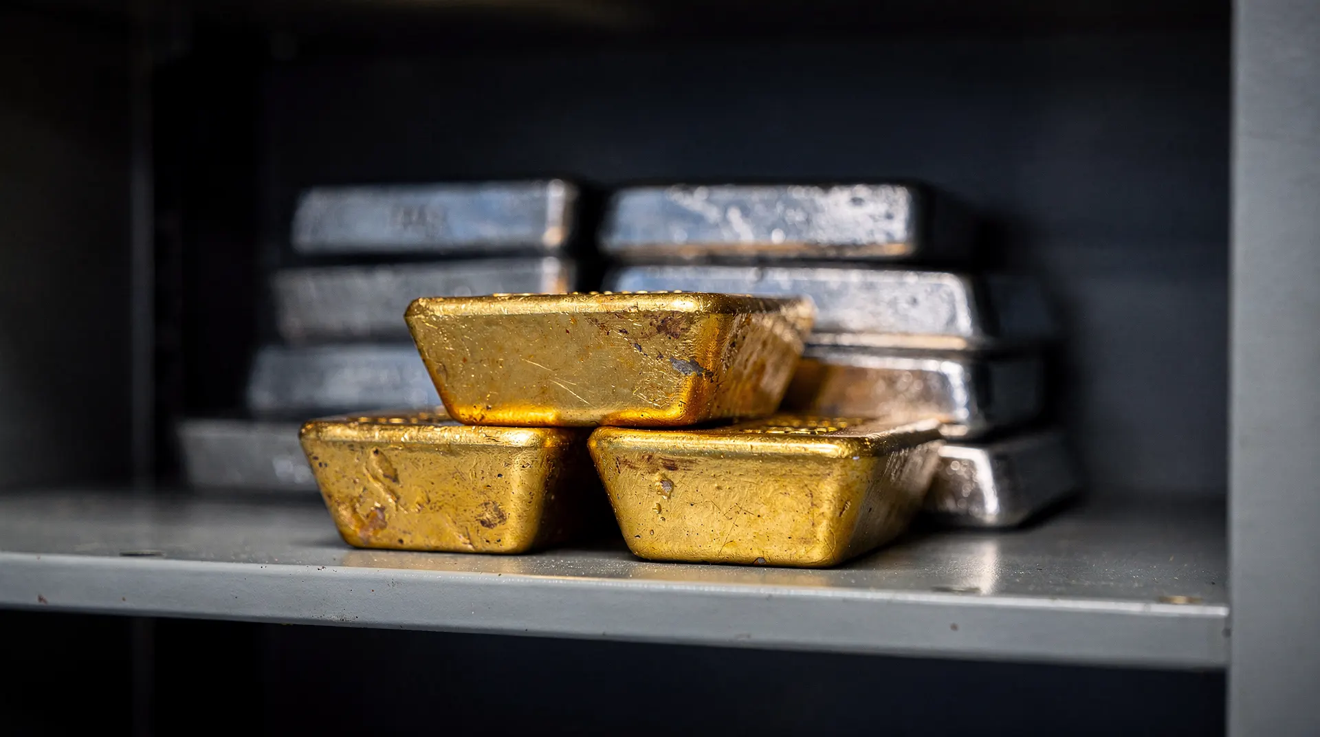 Unmarked gold bars sit in focus on a vault shelf, with silver ingots slightly behind them.