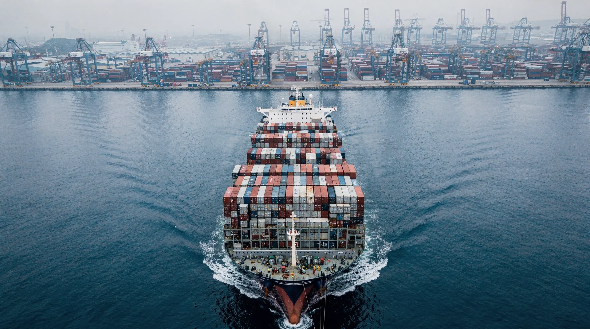 A container ship approaches a harbor terminal with unmarked containers and cranes.