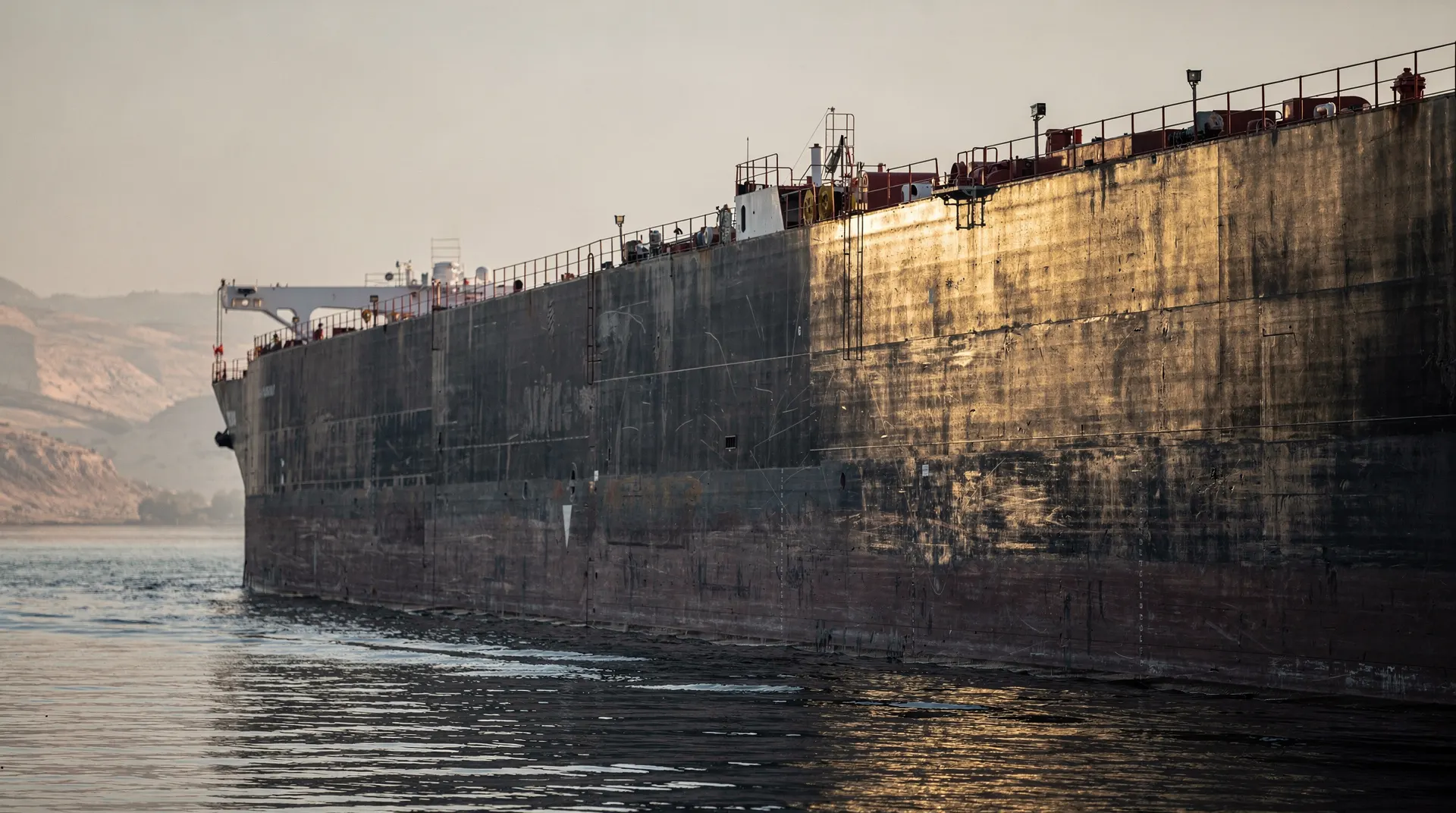 An unmarked oil tanker passes between hazy headlands across calm water.