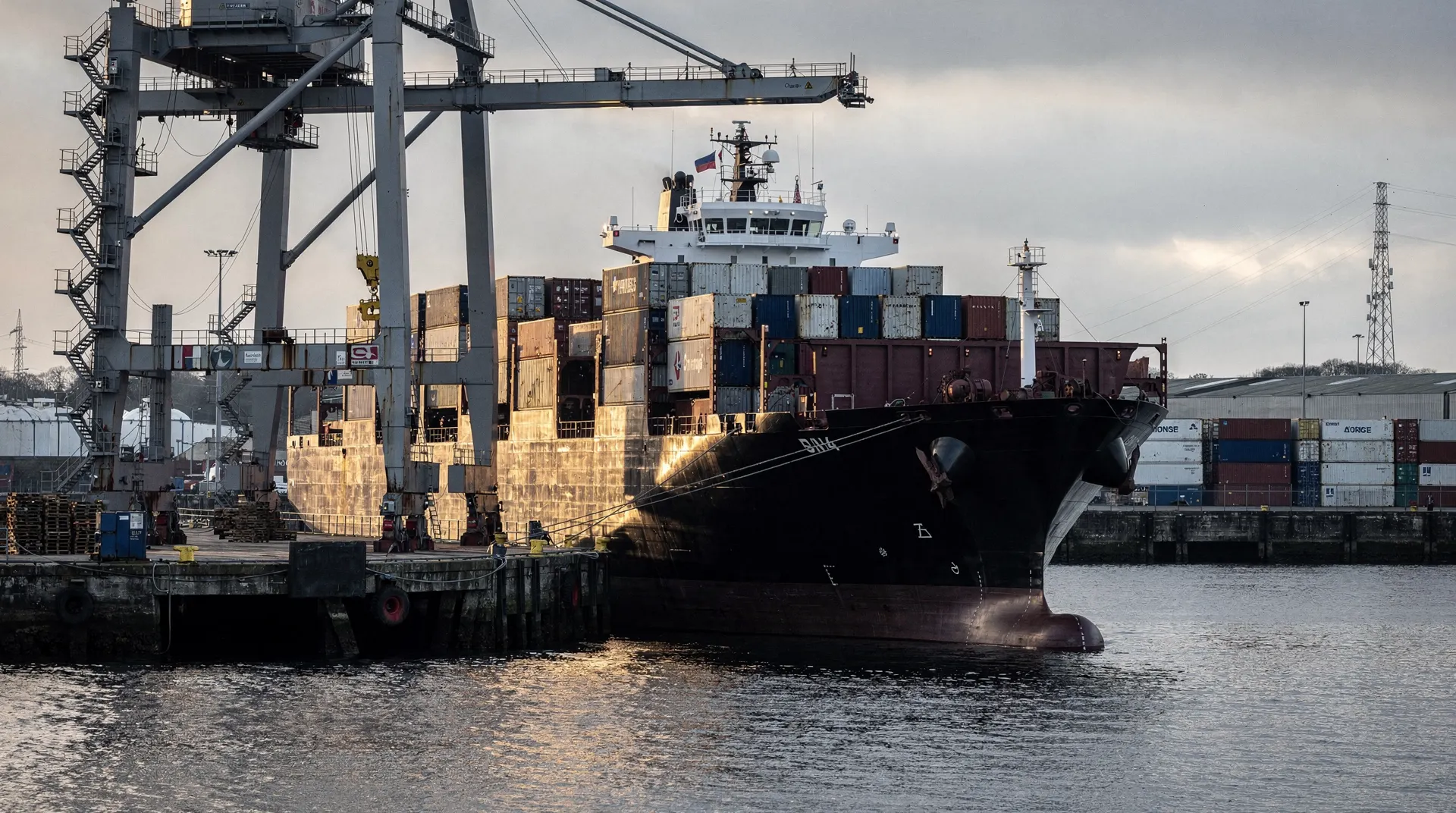An unmarked container ship sits beside cargo cranes and stacked containers at a port.