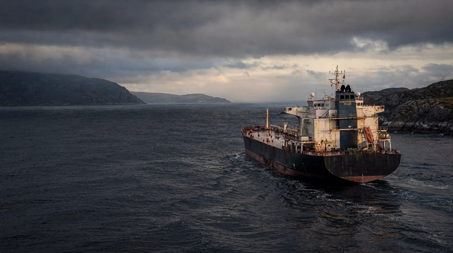 An unmarked LPG carrier crosses dark water beneath heavy clouds, with a hazy coastline in the distance.