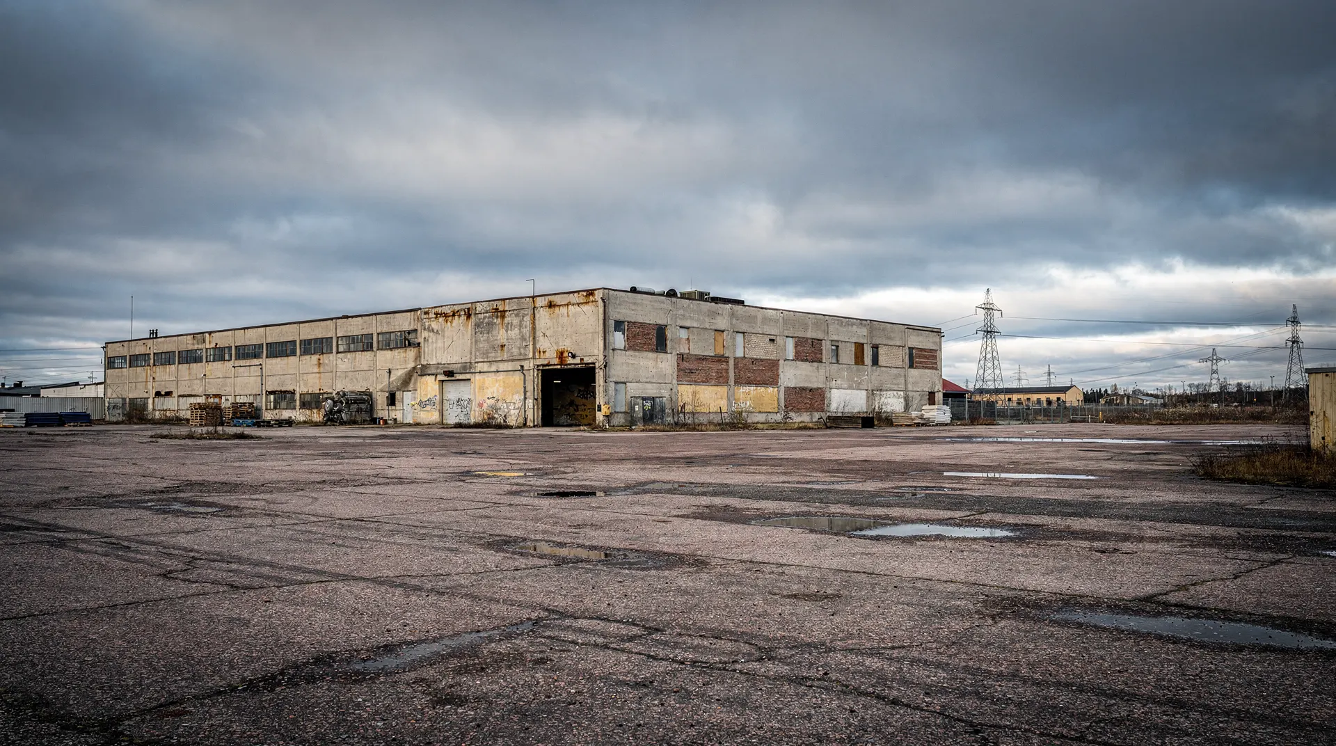 A weathered former paper mill faces an empty yard, with power pylons in the distance.