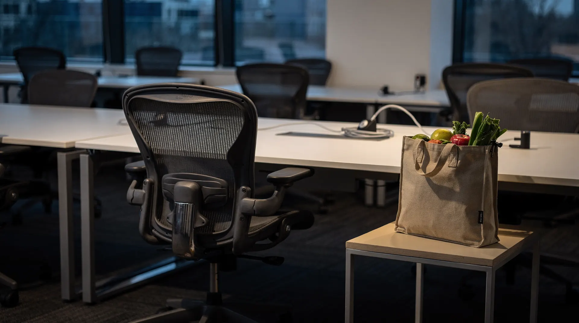 An empty office chair faces vacant workstations, with a grocery tote on a side table.