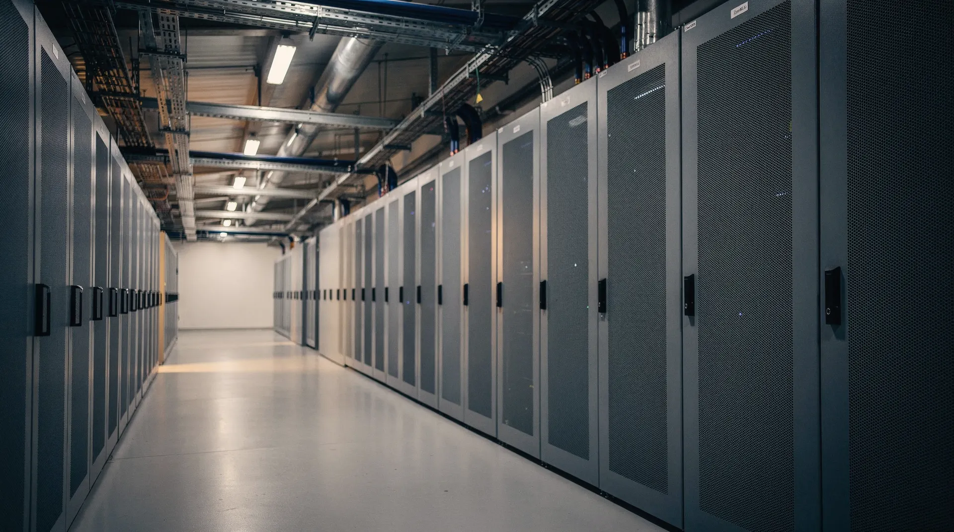 Unmarked server cabinets line a data-center aisle beneath overhead cooling pipes.