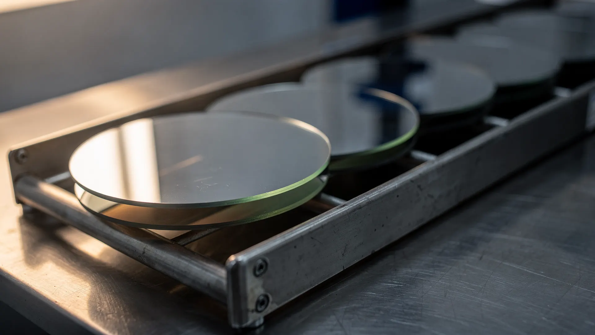 A brightly lit silicon wafer stands in a cleanroom carrier as other wafers recede into shadow.