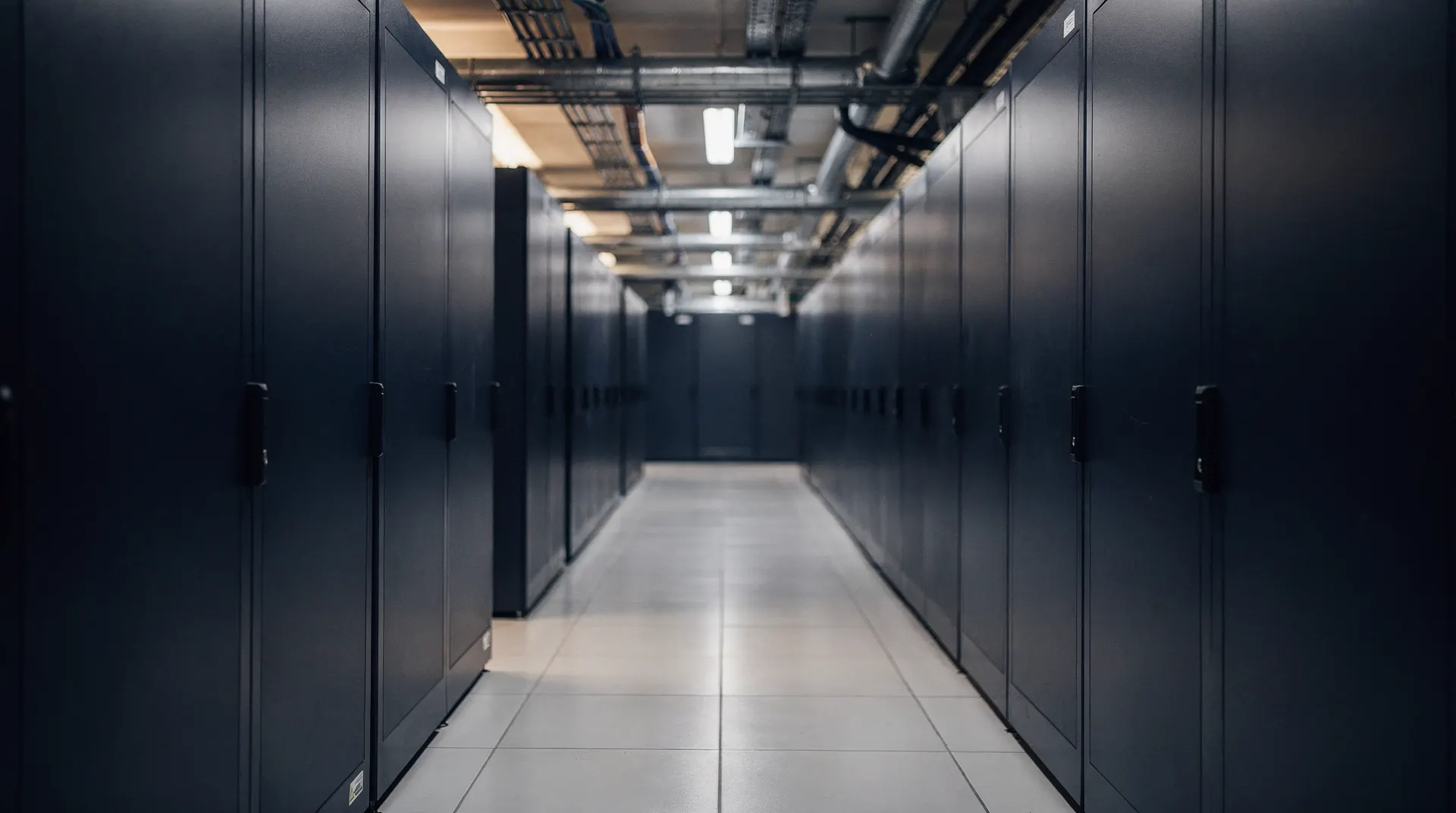 Closed server cabinets line a data-center aisle beneath cooling pipes.