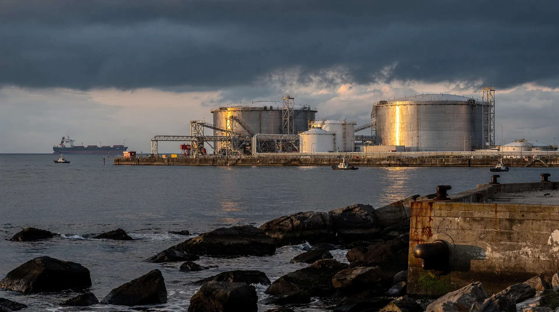 An oil tanker crosses a narrow channel beneath dark clouds, with calm water in the foreground.